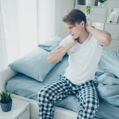 Junger Mann sitzt auf dem Bett, reibt sich den Nacken. Er hat nackenschmerzen nach dem schlafen.