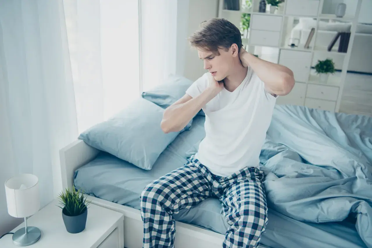 Junger Mann sitzt auf dem Bett, reibt sich den Nacken. Er hat nackenschmerzen nach dem schlafen.